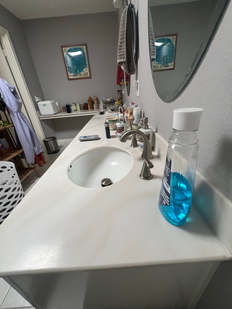 a stunningly clean midcentury/outdated double sink bathroom with oval mirrors, a collection of decorative tiki mugs along its wrap around counter, and assorted normal toilette instruments 