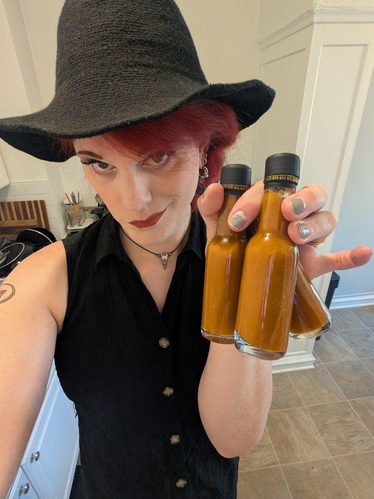 A selfie of me wearing a witch hat and holding three bottles of homemade hot sauce.