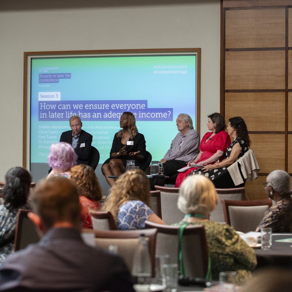A panel of two men and three women are sat on a stage. Behind them is a screen which reads Session 2: How can we ensure everyone in later life has an adequate income? In front of the stage are tables filled with different people watching the panel discussion.