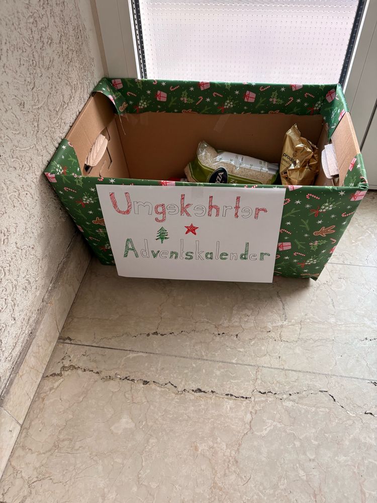 Pappkarton mit Geschenkpapier beklebt im Treppenhaus. Er trägt die Aufschrift „Umgekehrter Adventskalender“, darin liegen verpackte Lebensmittel.