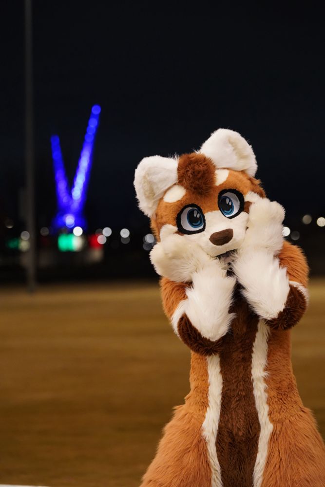 Fursuiter posing in foreground with Oklahoma City Scissortail bridge sculpture in background. Sculpture is illuminated as the picture was taken during night hours. 
