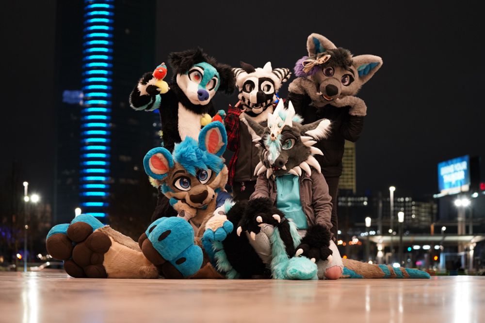 A group of fursuiters pose on an outdoor stage with a city skyline in the background during night hours. 