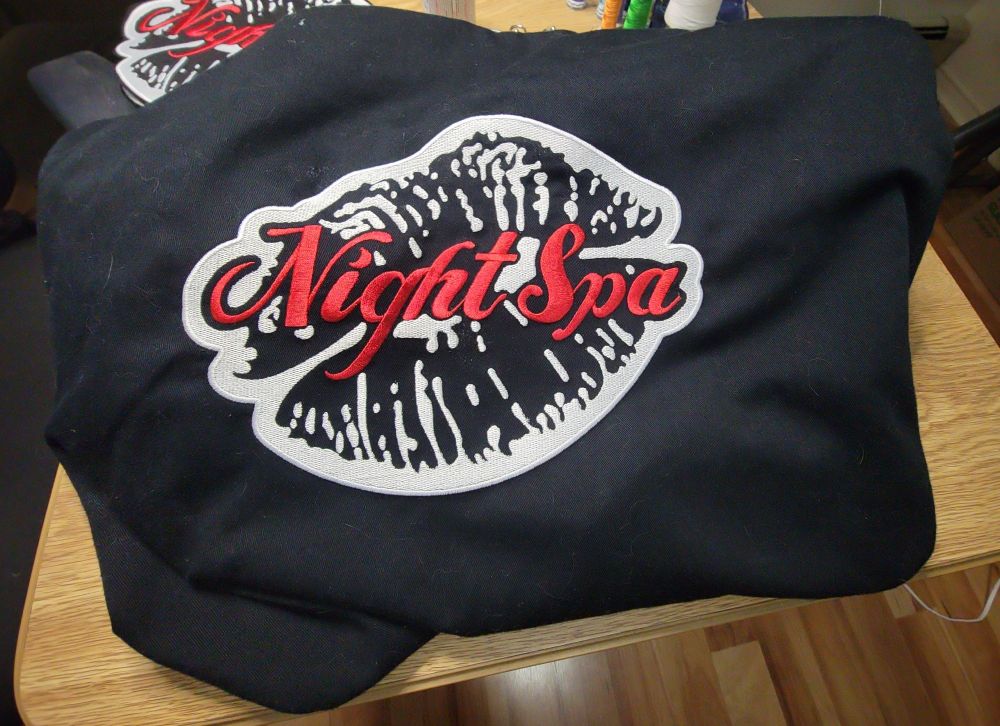 The back of a large black button-up shirt with a logo embroidered on it. The logo looks like lipstick leftover from a kiss with "Night Spa" overtop in red cursive