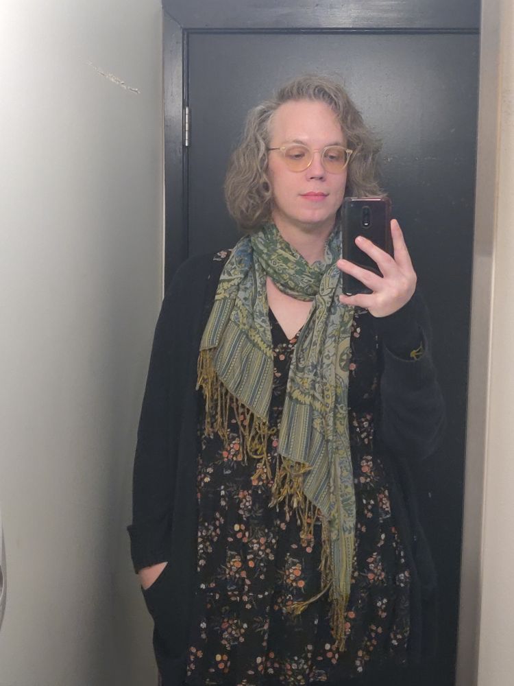 A woman in a floral print dress, black long cardigan, and green patterned scarf.