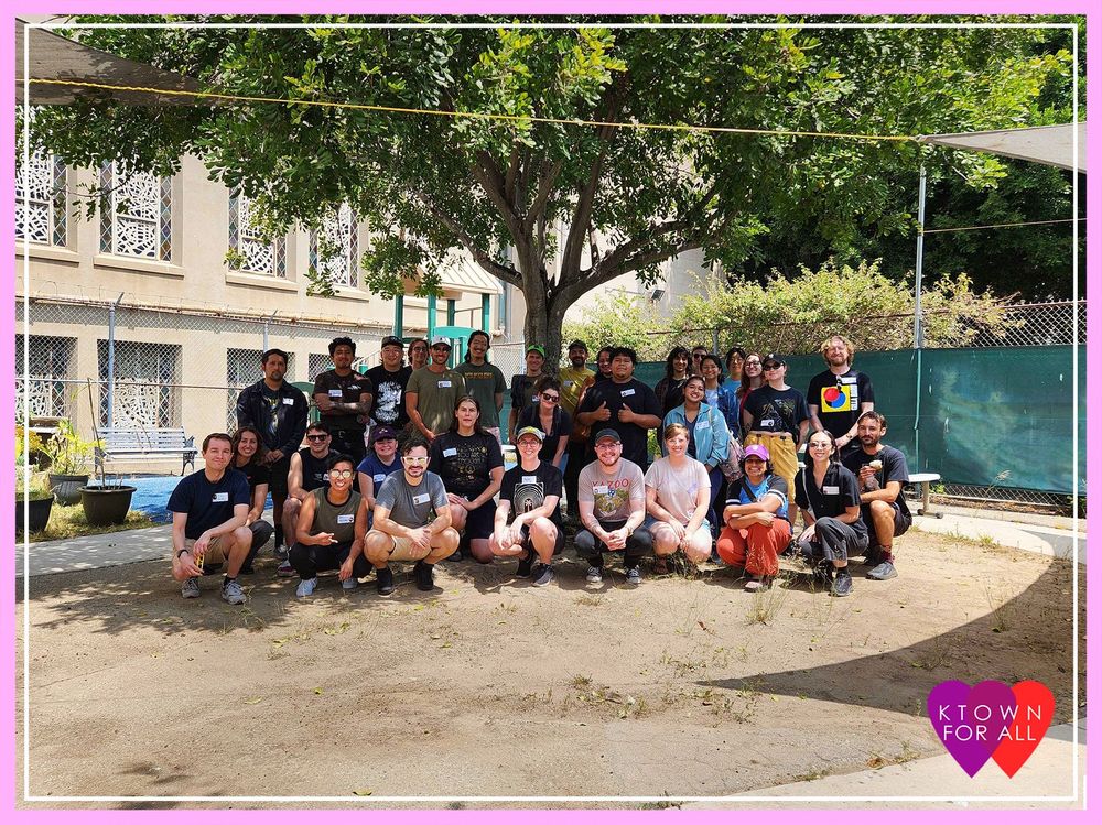 Group of around 20 people bunched in front of a tree, all smiling. There is a building and more trees behind the group of people. The picture is bordered with a thick light pink and a thin white line. There is an icon of overlapping purple and red heart with “KTOWN FOR ALL” in white letters centered on the overlapping hearts on the right bottom corner of the photo. 