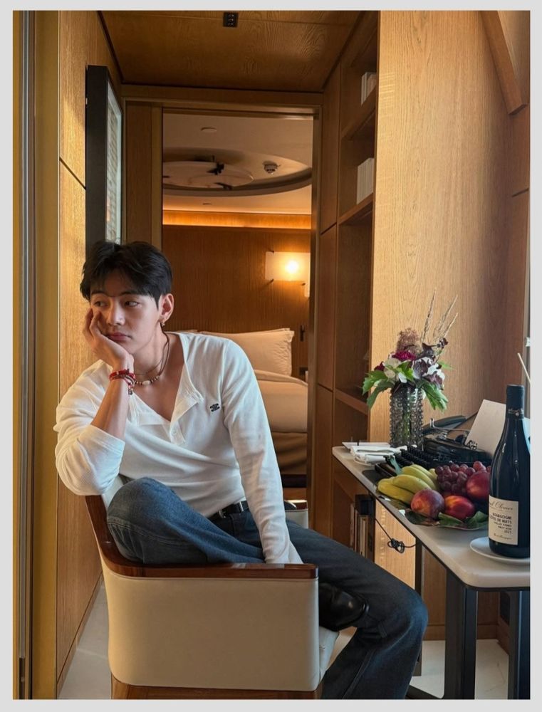 kim taehyung of bts poses pensively in a hotel chair looking offscreen. there is a table beside him with a bottle of wine and a plate of fruit. 