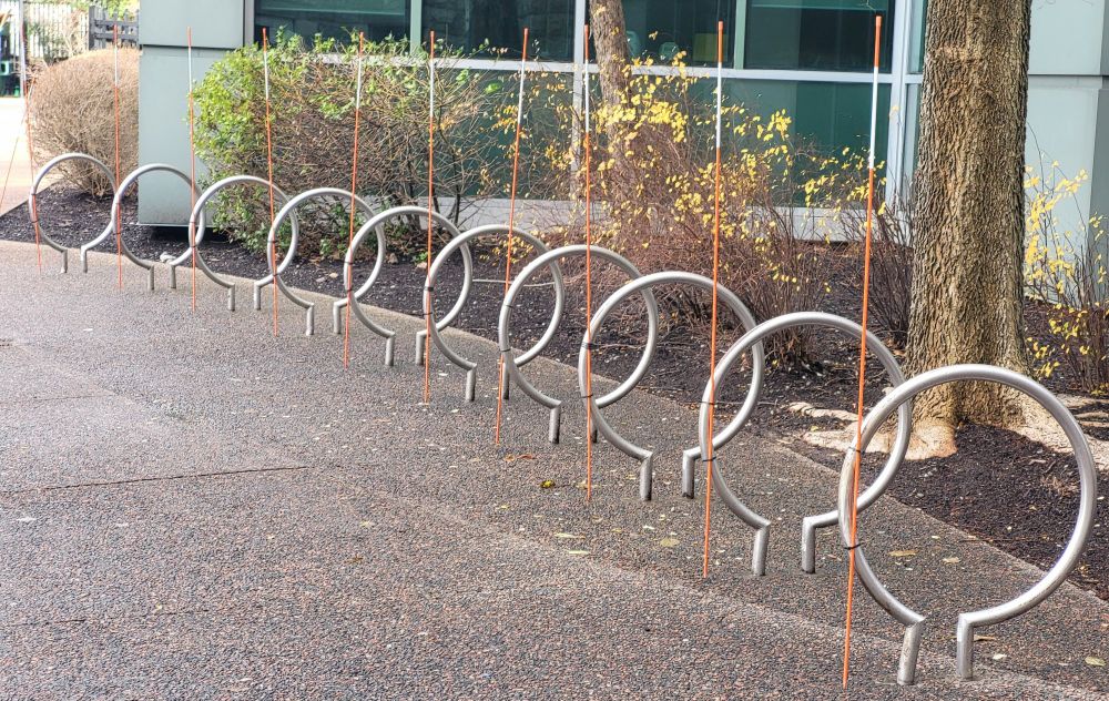 A series of round bicycle locking frames arcs to the left along a path. Each of the frames has an orange snow-plow warning stick attached to it that points directly upward.