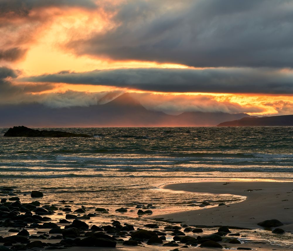 The sun rises over distant mountains giving a yellow cast to the sky that peeps through the hard blue cloud cover. Sea mist is lit bright red by the off-axis sun, while waves beach towards the viewer.
