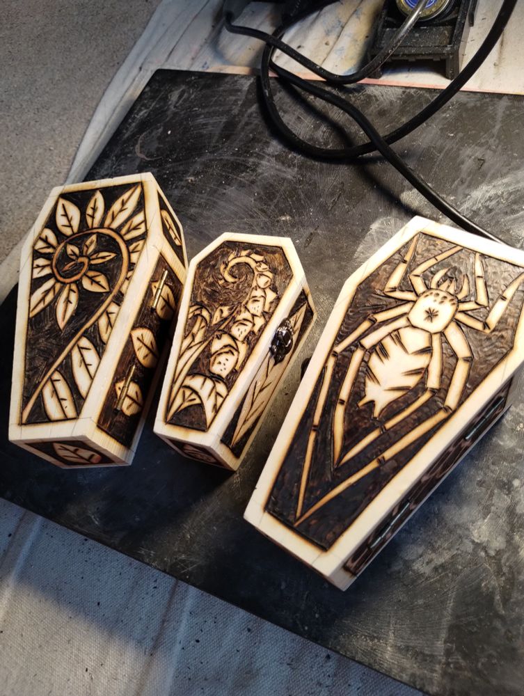 Three mostly finished wood boxes with wood burned designs. A curling fern. A foxglove. And a spider 