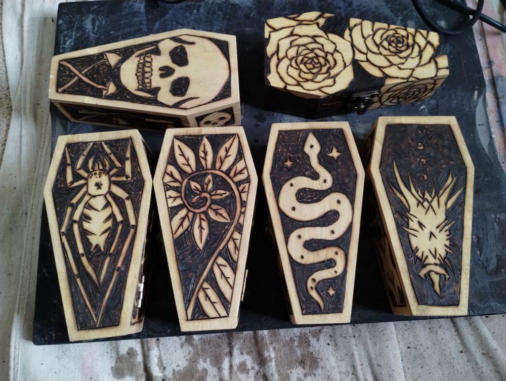 A set of mini wood coffins with pyrography designs on them- a skull with mushrooms, roses, spider, fern, snake, and thorn apple pod.