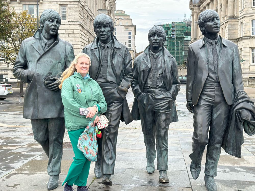 My wife, a white woman with blonde hair, smiling and wearing green jacket and green pants, standing in front of statues of the four members of The Beatles in Liverpool, UK. 