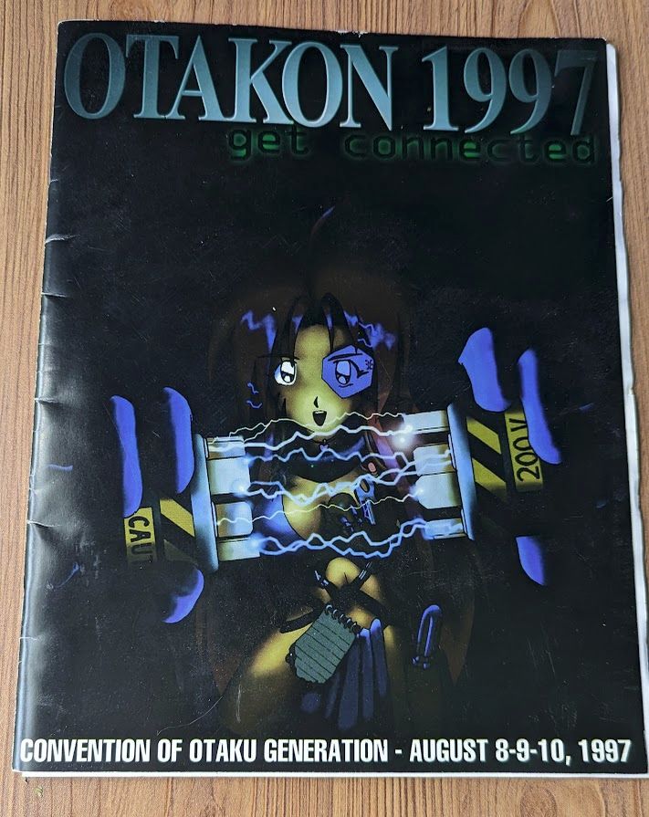 The Otakon 1997 con book. It has an old-fashioned looking anime girl in some kind of fantasy bikini on the cover, holding up some sort of electronic plugs that are crackling with electricity, in front of a black background. It reads "OTAKON 1997 get connected" along the top and "CONVENTION OF OTAKU GENERATION- AUGUST 8-9-10, 1997" along the bottom.