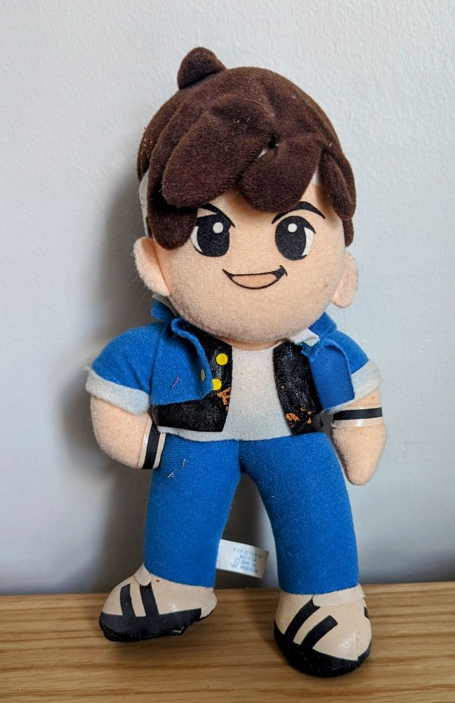 A plush toy of Shingo from King of Fighters, like one you'd get from a UFO catcher. His white shoes are a little faded, and he has some brown vinyl pieces on him that fell off of another toy. The toy is old-fashioned but very cute.