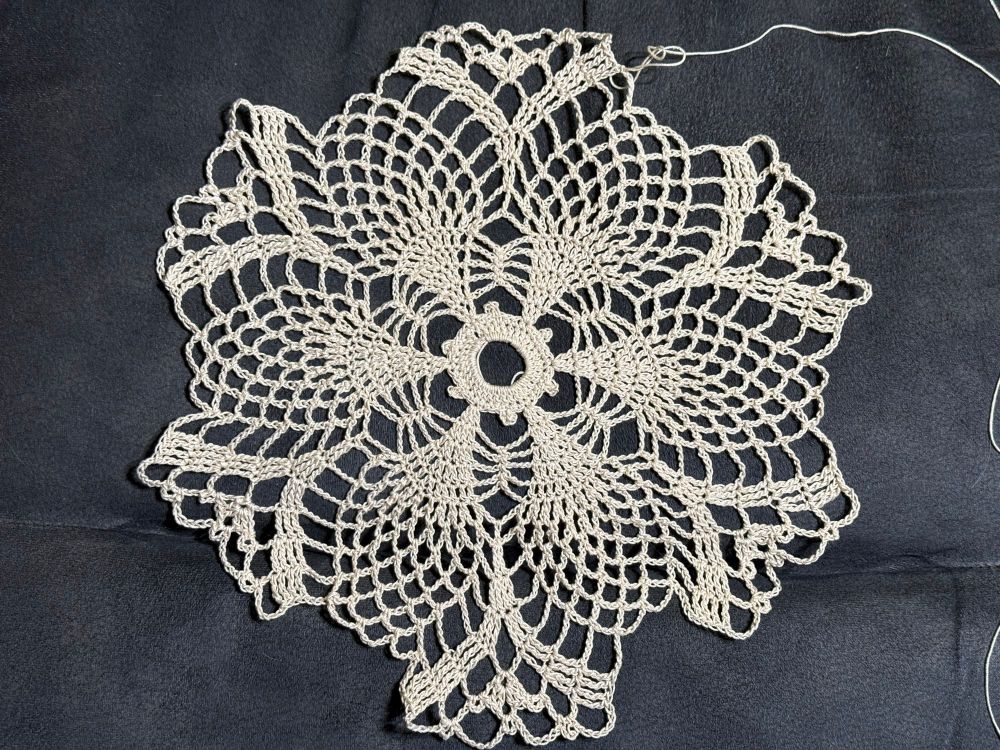 An off white crochet doily showing more flower petals on a dark background. 