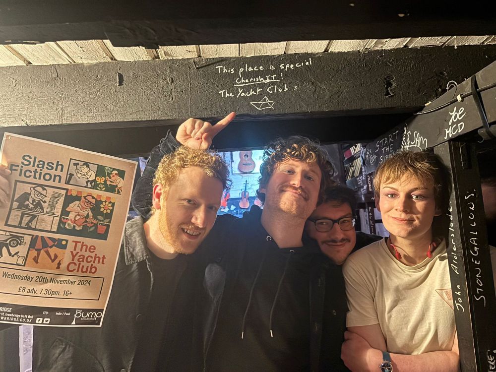 From left to right Ali, Marcus, Tom and Reverie under the beam it signed. 
“This place is special, cherish it. The Yacht Club” 

Ali is holding up a poster of the gig, Marcus is pointing at the message. Tom’s head is poked in between Marcus and Reverie’s. Reverie has its arms folded and it’s smiling. She has been crying a lot because they got to see trans teenagers dancing to their music today. 