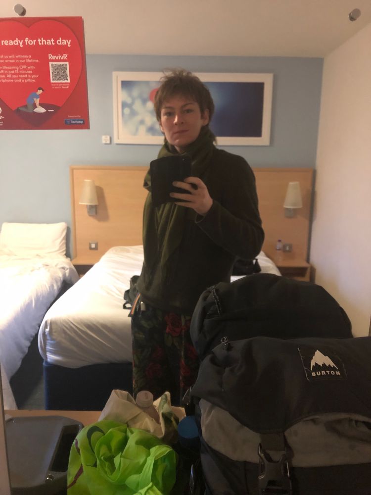 A hotel mirror selfie of Reverie in her floral trousers, green jumper and scarf. In the foreground are various bags