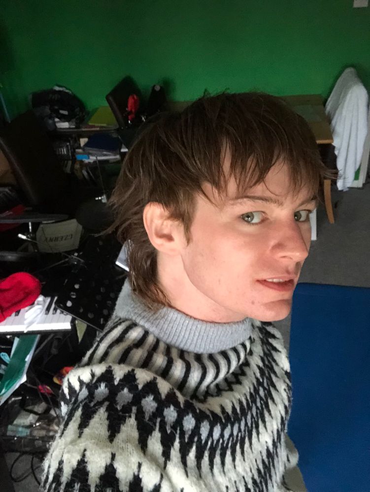 Reverie in her living room with big white jumper on that has like a half-diamond pattern thing going on. 

It’s hair looks messy (good) 