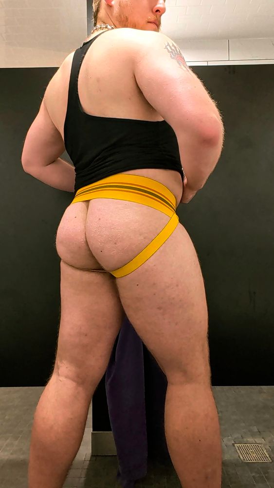Me in the gym bathroom stall, only wearing black stringer and yellow jock, back to camera and peering over shoulder, glutes and thighs looking very pumped