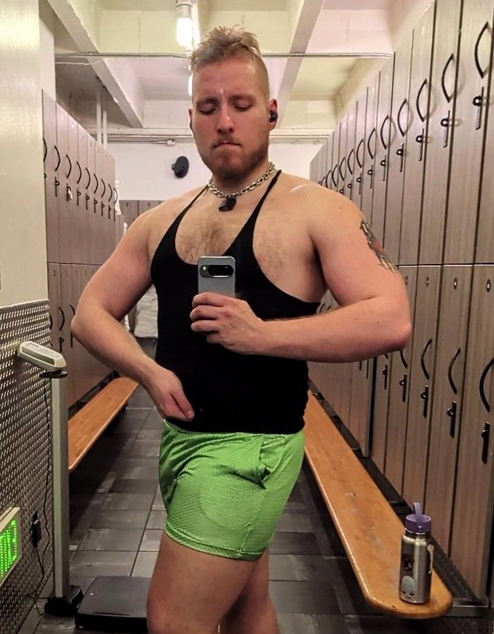 Me in gym locker room mirror, black stringer and green shorts, twisting body to one side and flexing chest