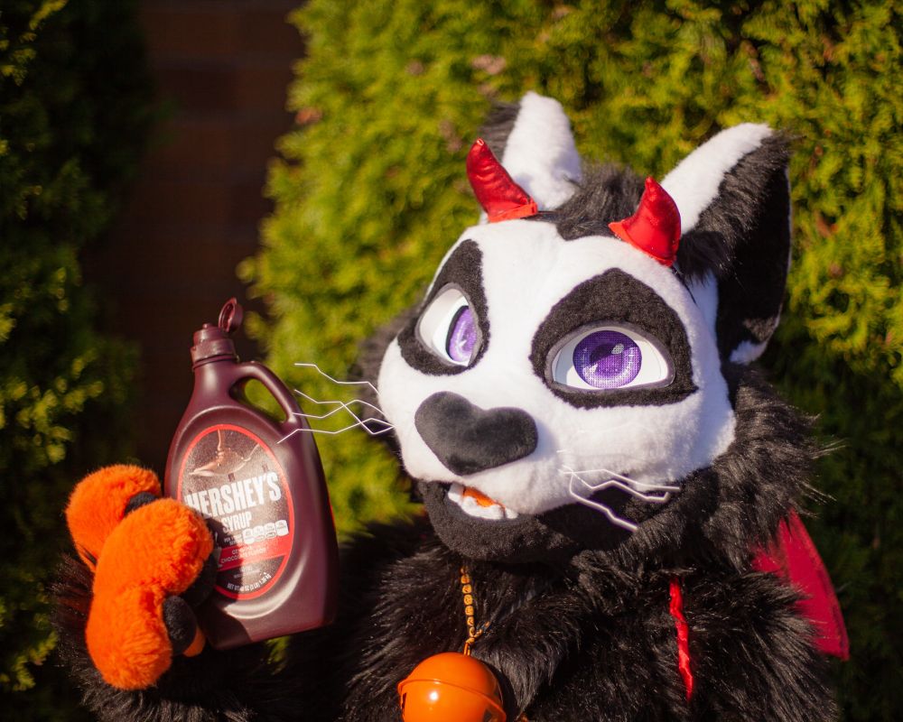 A photo of a halloween themed cat fursuiter in a silly devil costume holding up the bottle of Hershey's syrup he uses as a water bottle
