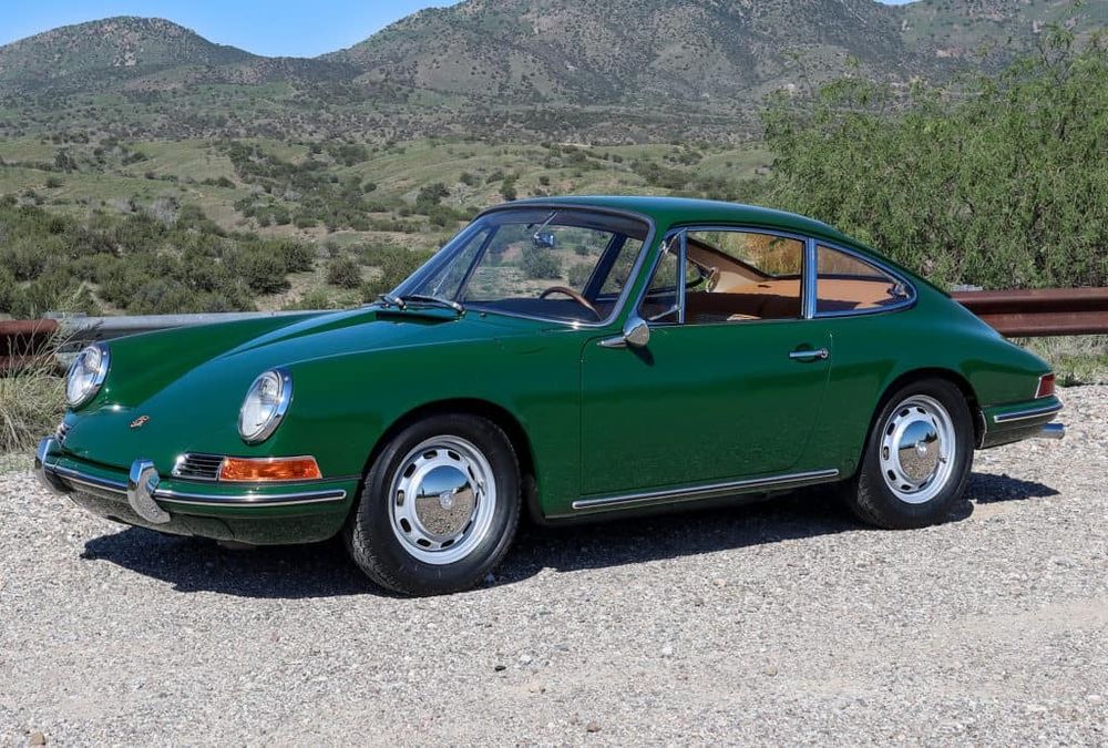 The image features a green classic Porsche car, likely a vintage model with its distinct round headlights and sleek, timeless design. The car is parked on a gravel path with a scenic backdrop of green hills and clear blue skies. The shiny chrome detailing and well-preserved exterior highlight its classic appeal, contrasting beautifully with the natural landscape.