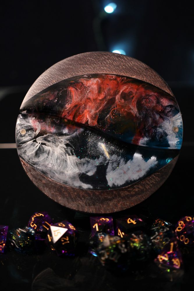 Round sycamore dice box stained dark brown with a large oval cutout filled with epoxy that looks like a small figure holding up a blazing sword in the path of a billowing red black and white cloud.