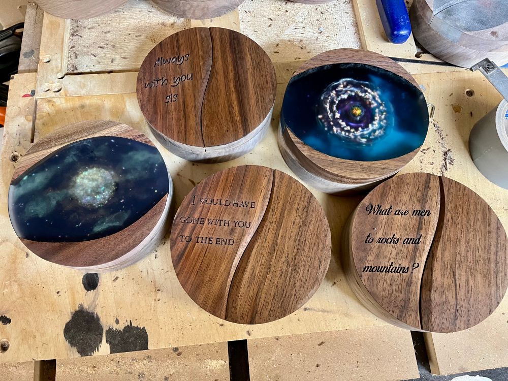 Five round dice boxes made from walnut. Two have epoxy incorporated into the top. One looks like a ringed planet or some kind of space phenomena. The other an ethereal moon is partially obscured by iridescent green clouds. The others don’t have epoxy but have quotes etched into the tops. One says Always With You Sis, another says What are men to rocks and mountains, the last saying I would have gone with you until the end