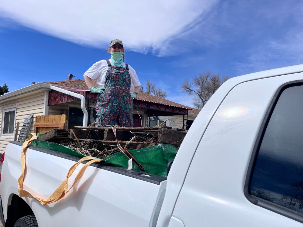 Yours truly, a white woman in colorful overalls, kneeling atop the pile of weeds, weighted down with pallets, in the bed of a full size truck