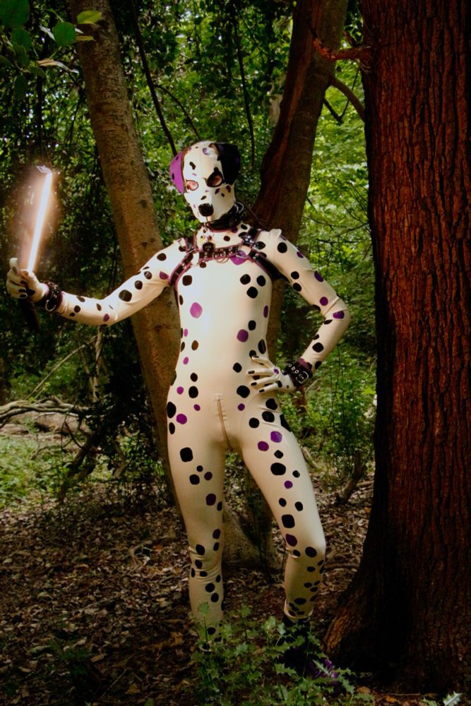 Dalmatian Styra fully covered in white latex with black and purple spots, is stood beside a tree with their left leg bent slightly holding a light strip in their right paw.

The photo is very striking and moody!