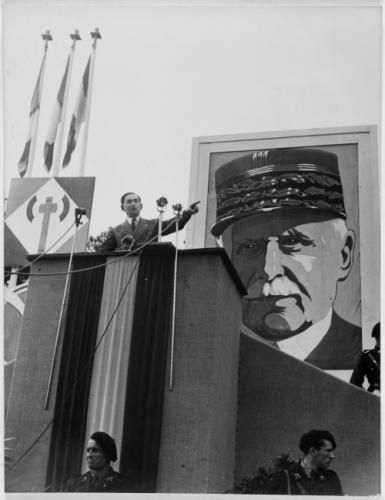 Speaker standing at huge podium probably addressing a crowd in Vichy France about the glories of collaborating with the Nazis, surrounded by guards, while a giant picture of Marechal Petain looms behind him.