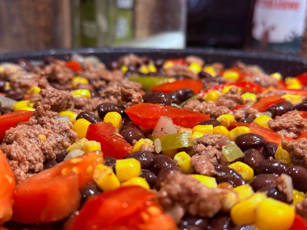A pan of simmering tomatoes, ground beef, black beans, corn, onion, and jalapenos.