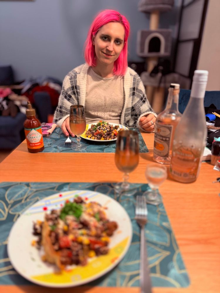 @kanba.gay smiling adorably over two plates of the mexicasserole, with a bottle of hot sauce and two glasses of wine on the table.