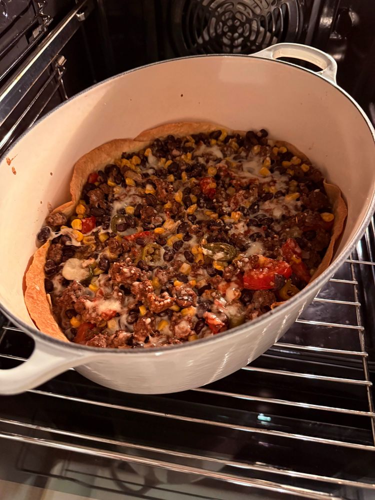 A casserole pot lined with crispy corn tortillas and filled with ground beef, tomatoes, jalapenos, corn, black beans, onion, cheese, and sour cream coming out of the oven, looking beautiful.