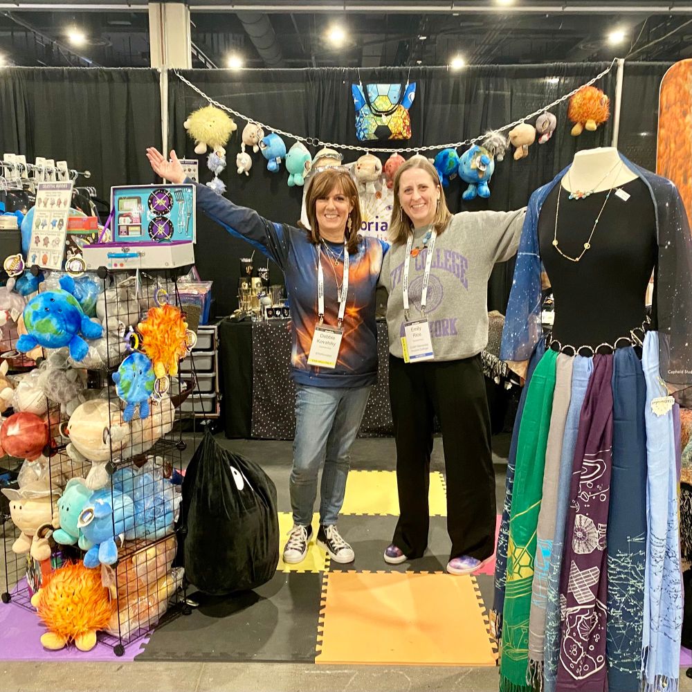 Debbie and Emily posing inside the BOOTHtique in the AAS conference exhibit hall, with plush planets, mission scarves, and jewelry on display (plus lots more!)