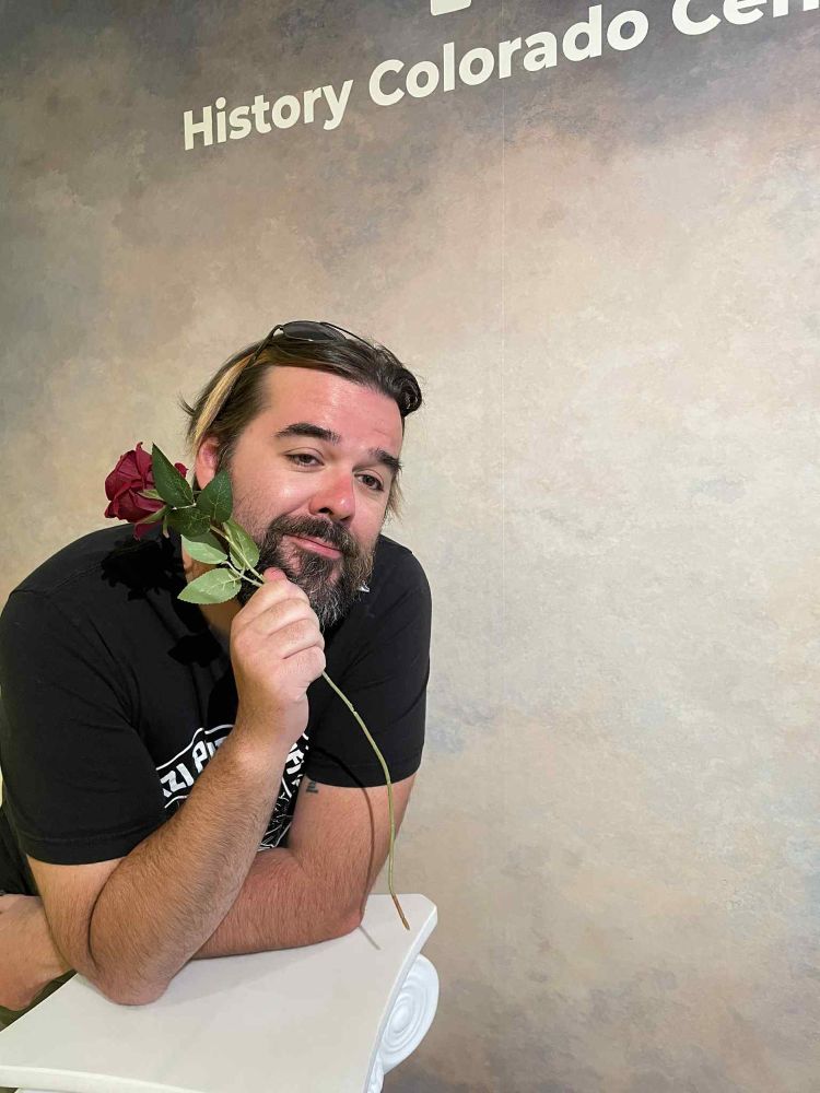 A bearded person in a black shirt with a rose in a glamor shots pose