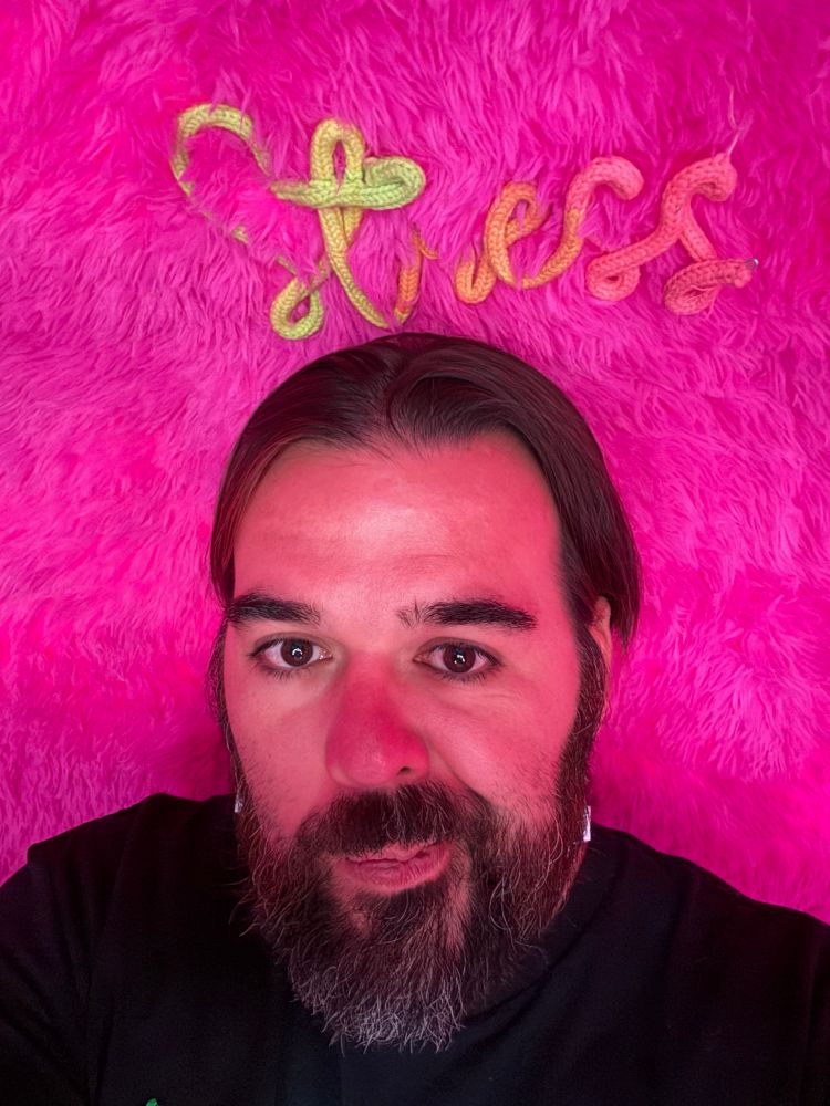 A bearded person in a black shirt against a pink background. The word stress in cursive is stitched in the wall.