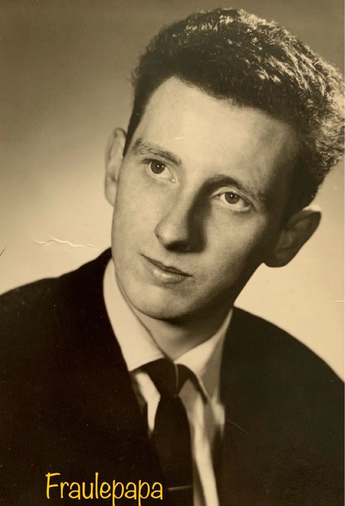 Ein Sepia Bild von einem jungen Mann mit schwarzer Anzugjacke, weißem Hemd und schwarzer Krawatte. Die dunklen kurzen Haare lockig. Perspektive vom Betrachter: Der Kopf ist nach rechts geneigt. Die Ohren stehen leicht im Wind. Der Blick geht nach links oben, etwas verklärt. Links unten steht Fraulepapa