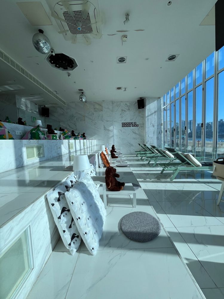 Ein helles, modernes Interieur mit Marmorboden. Liegestühle sind in der Nähe großer Fenster angeordnet, mit Plüschtieren und dekorativen Decken. Die Decke hat dekorative Elemente, darunter eine Discokugel. Natürliches Licht erfüllt den Raum und schafft eine entspannte Atmosphäre.