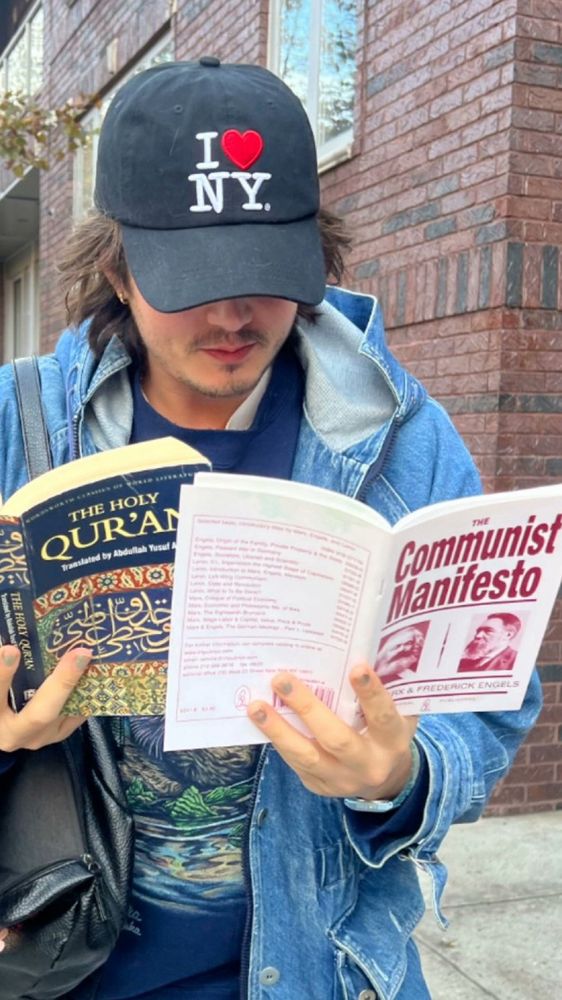 a young white guy in a jean jacket is reading both the qur’an and the communist manifesto at the same time while wearing a “I ❤️ NY” ballcap.