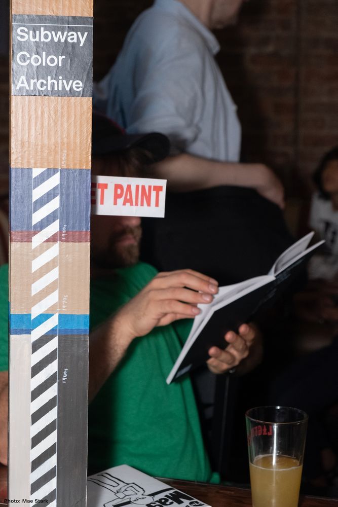 A model subway poll and wet paint sign obscure a man reading. Photo by Mae Stark.