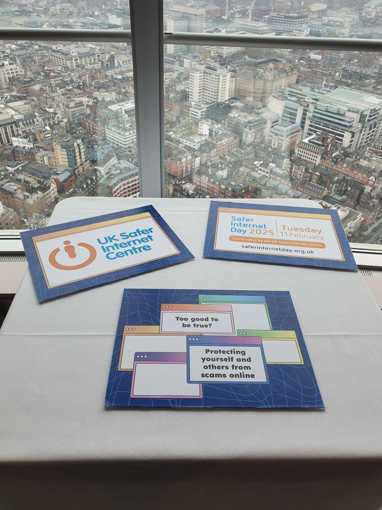 Aerial view of London through the window. Table in front, holding signs that say "UK Safer Internet Centre", "Too good to be true? Protecting yourself and others from scams online"