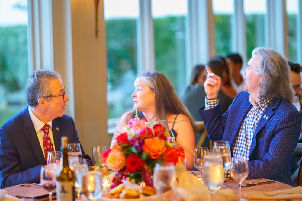 Three people engaging in a conversation at a dining table adorned with a vibrant floral centerpiece, in a room with large windows.