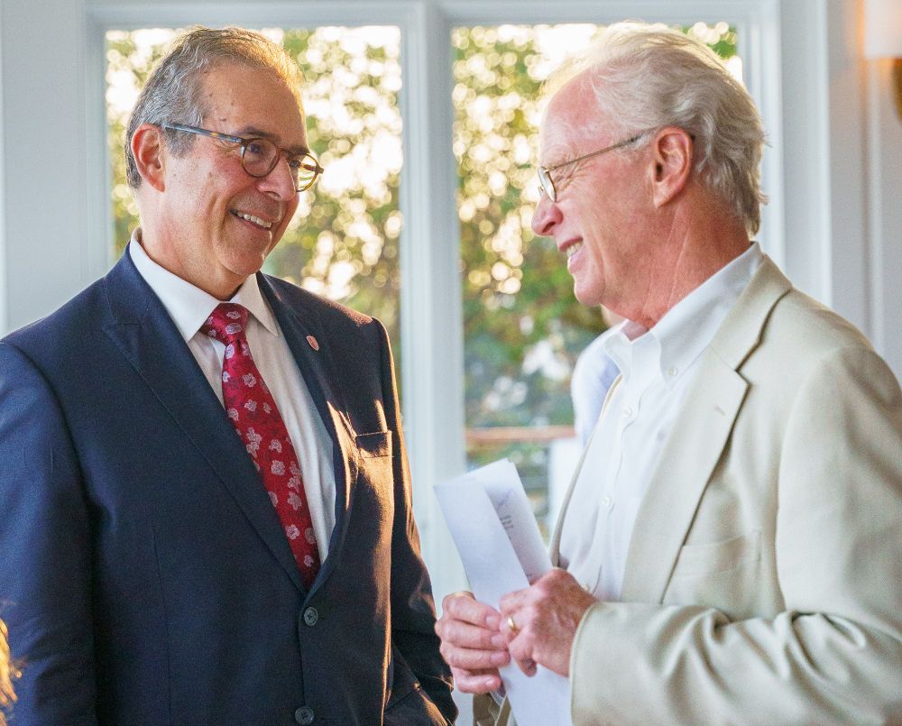 Two individuals in formal attire engaging in a friendly conversation near a window in a sunlit room.