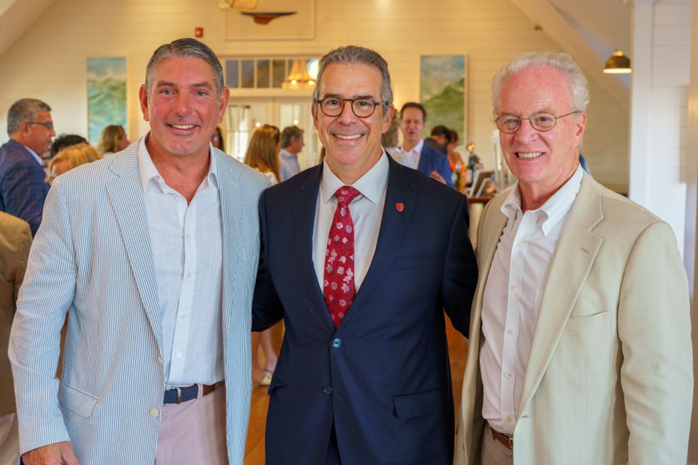 Three individuals smiling at a social gathering, with two dressed in casual blazers and one in a suit with a red tie. They are inside a room with artwork and other people in the background.