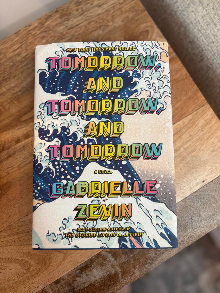 Photo of the book cover for 'Tomorrow, and Tomorrow, and Tomorrow' by Gabrielle Zevin, placed on a wooden surface.