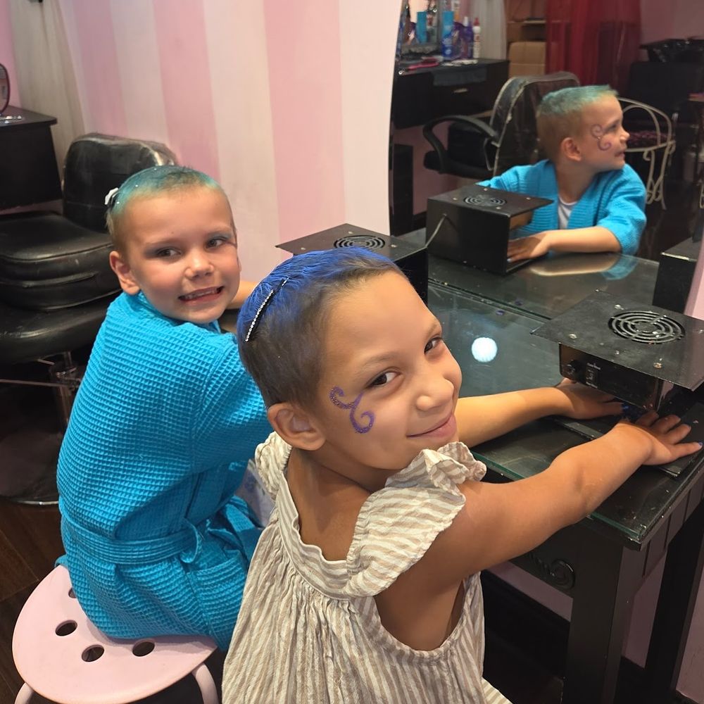 Two children sitting at a salon station, each wearing a salon cape and sporting a fresh hairstyle with a design painted on their cheeks, smiling at the camera.
