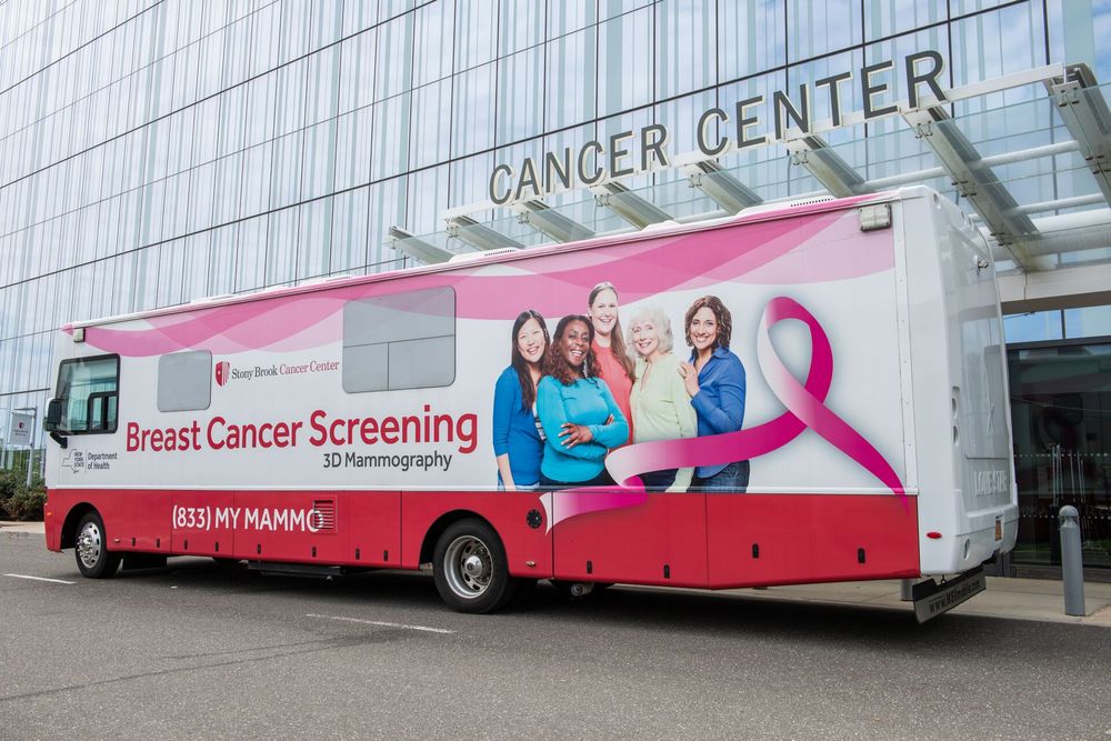 A mobile breast cancer screening bus featuring an advertisement for 3D mammography services parked outside a building labeled "Cancer Center". The bus is decorated with a large pink ribbon and images of smiling individuals.