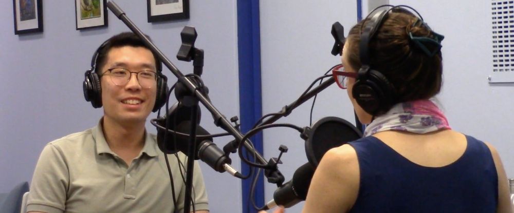 Two people are sitting in a radio studio, facing each other with microphones in front of them, smiling as they engage in a conversation. They are wearing headphones.