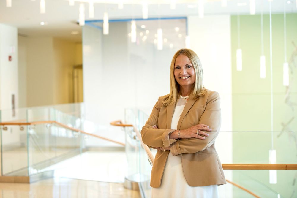 Professional individual standing with arms crossed in a modern office lobby, smiling confidently.