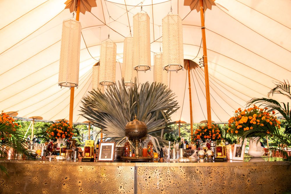 An elegant event setup under a tent with lantern-style lighting hanging from above. The decor features a lush arrangement of tropical plants and vibrant orange flowers at the center of a long table adorned with various beverages and framed pictures.
